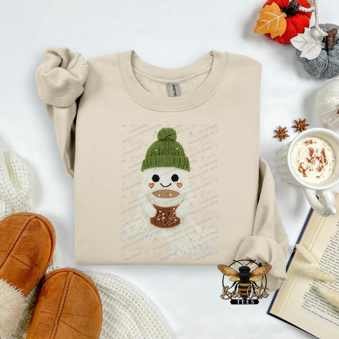 A folded beige long-sleeve shirt with a cartoon ghost graphic wearing a green beanie and brown scarf, styled in a flat lay with a hat, jeans, leaves, a book, and a cup on a white background.