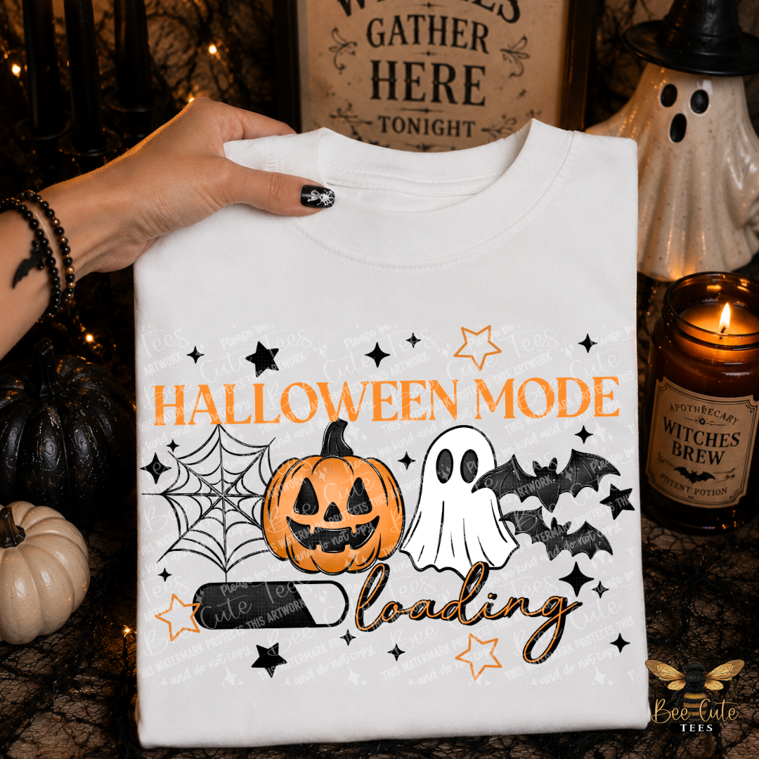 A white crewneck sweatshirt held up by a hand, featuring a Halloween-themed graphic print with a jack-o'-lantern, ghost, bats, spiderwebs, and the text 'HALLOWEEN MODE loading'. The background is a styled autumn scene with candles, a ceramic ghost, and pumpkins.