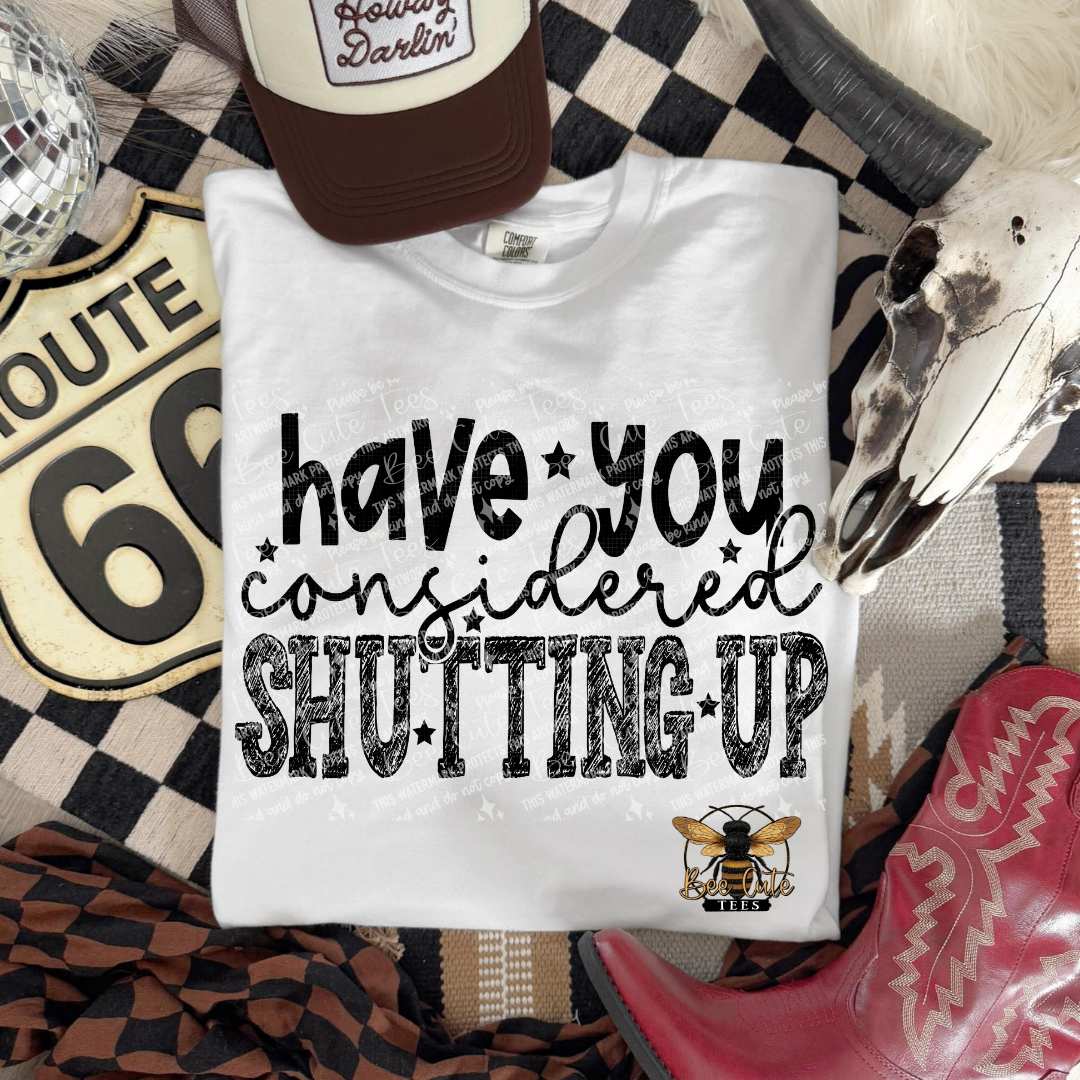 A white graphic t-shirt laid flat on a western-themed background with a checkered blanket, cowboy boots, a Route 66 sign, a hat, and a skull. The shirt features the text 'have you considered SHUTTING UP' in black distressed lettering with a bee logo below it.