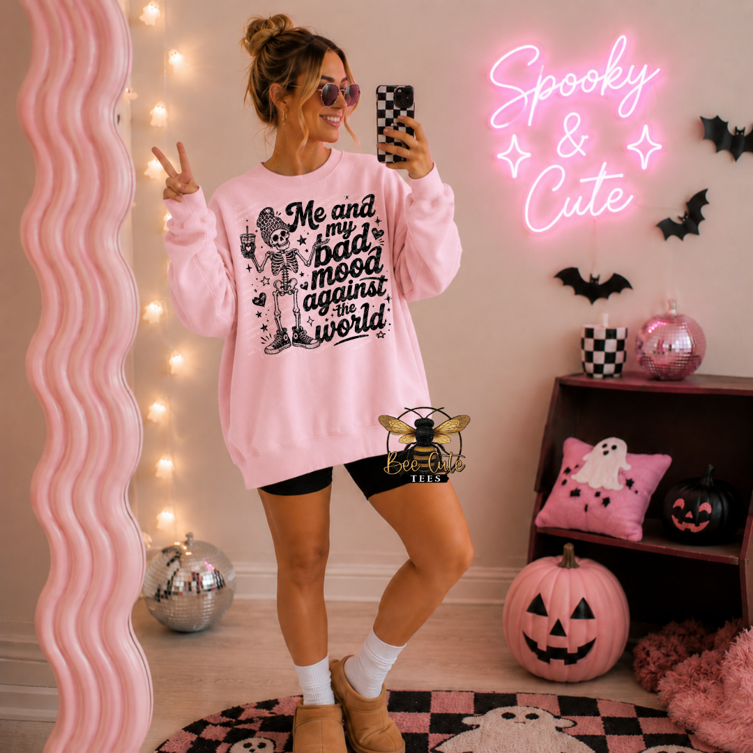 A woman takes a mirror selfie wearing a light pink crewneck sweatshirt with a black skeleton graphic and the text 'Me and my bad mood against the world'. The room is decorated for Halloween with a pink neon sign reading 'Spooky & Cute', a wavy pink mirror, pumpkins, and a checkered floor.
