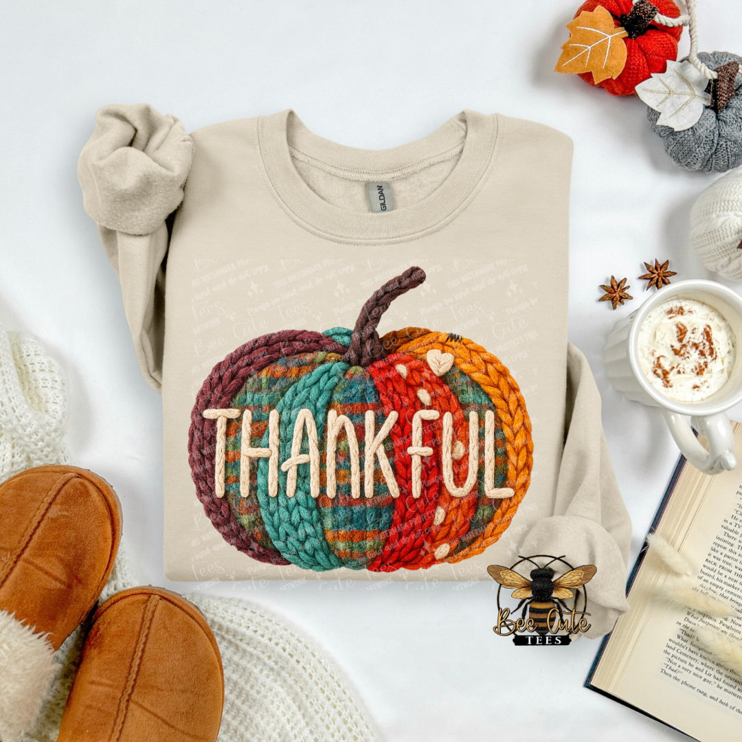 A beige crewneck sweatshirt laid flat on a white surface, featuring a large autumn-themed graphic of a knitted pumpkin with the word 'THANKFUL' across it. The sweatshirt is styled with cozy props including tan slippers, a mug of coffee with whipped cream, an open book, and fall leaves.