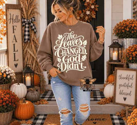 A woman wearing a taupe brown crewneck sweatshirt with a cream graphic that reads 'The Leaves Changed, God Didn't.' stands on a decorated fall porch with pumpkins, flowers, and autumn decorations.