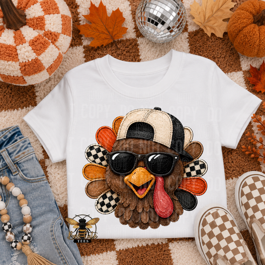 A white short-sleeve T-shirt laid flat on a fall-themed rug with pumpkins, leaves, a disco ball, jeans, beads, and checkerboard shoes around it. The shirt features a large turkey graphic wearing a beige-and-black cap and black sunglasses.