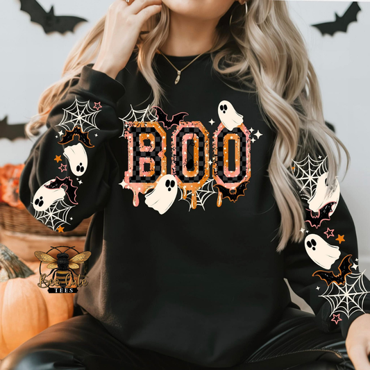 A woman wearing a black long-sleeve sweatshirt with a Halloween 'BOO' graphic on the chest and spiderwebs and ghosts printed on both sleeves. The background features a light gray wall with black bat cutouts, a pumpkin, and a decorative bee logo in the bottom-left corner.