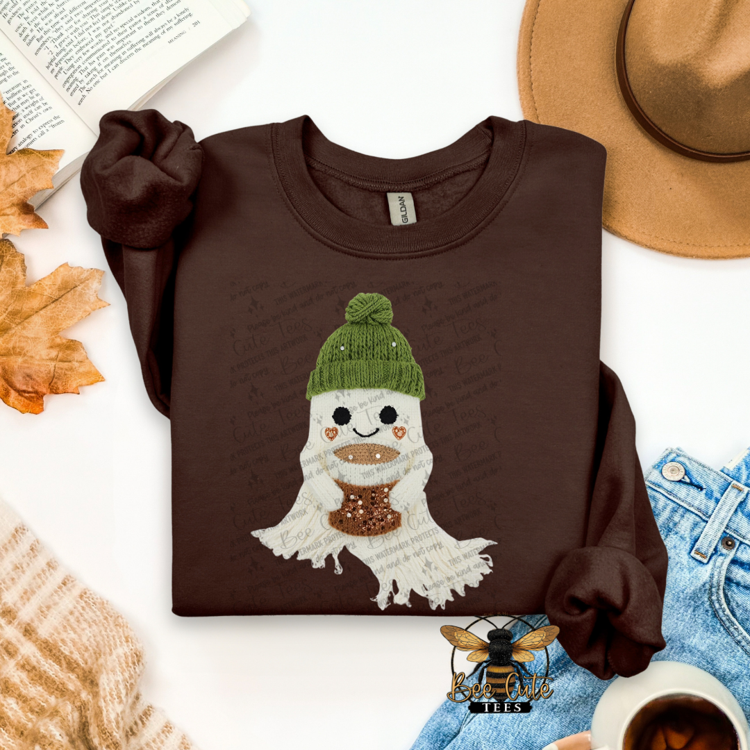 A folded brown long-sleeve shirt with a cartoon ghost graphic wearing a green beanie and brown scarf, styled in a flat lay with a hat, jeans, leaves, a book, and a cup on a white background.