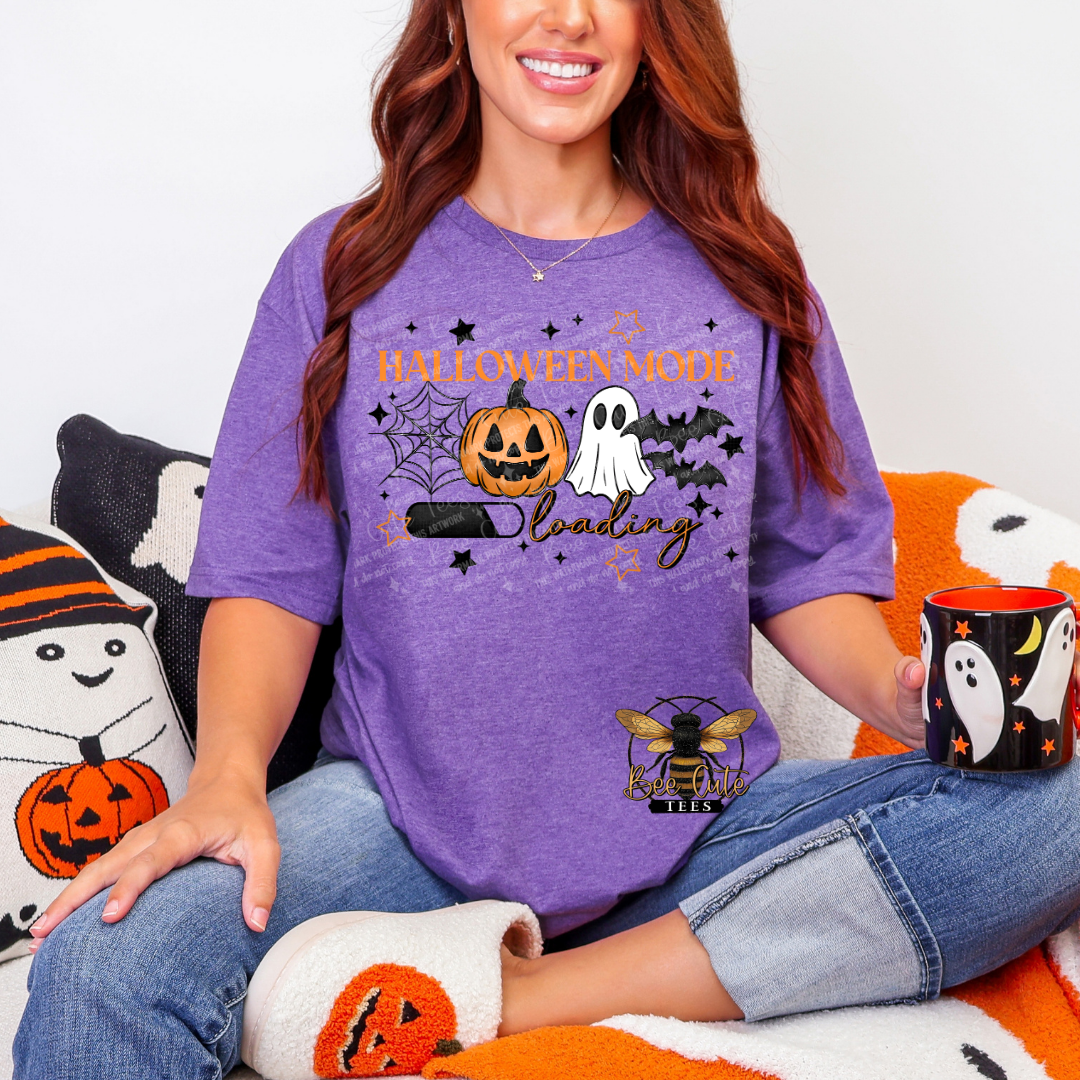 A woman wearing a purple heather graphic t-shirt with a Halloween-themed 'Halloween Mode Loading' design, sitting on a cozy blanket with Halloween pillows and a mug.