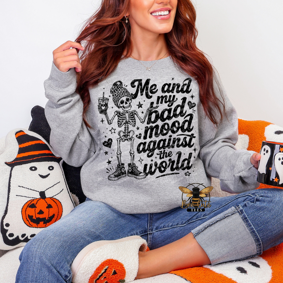 A woman wearing a light gray crewneck sweatshirt with a black graphic of a skeleton and the text 'Me and my bad mood against the world'. She is sitting on a white couch with Halloween-themed pillows and blankets.