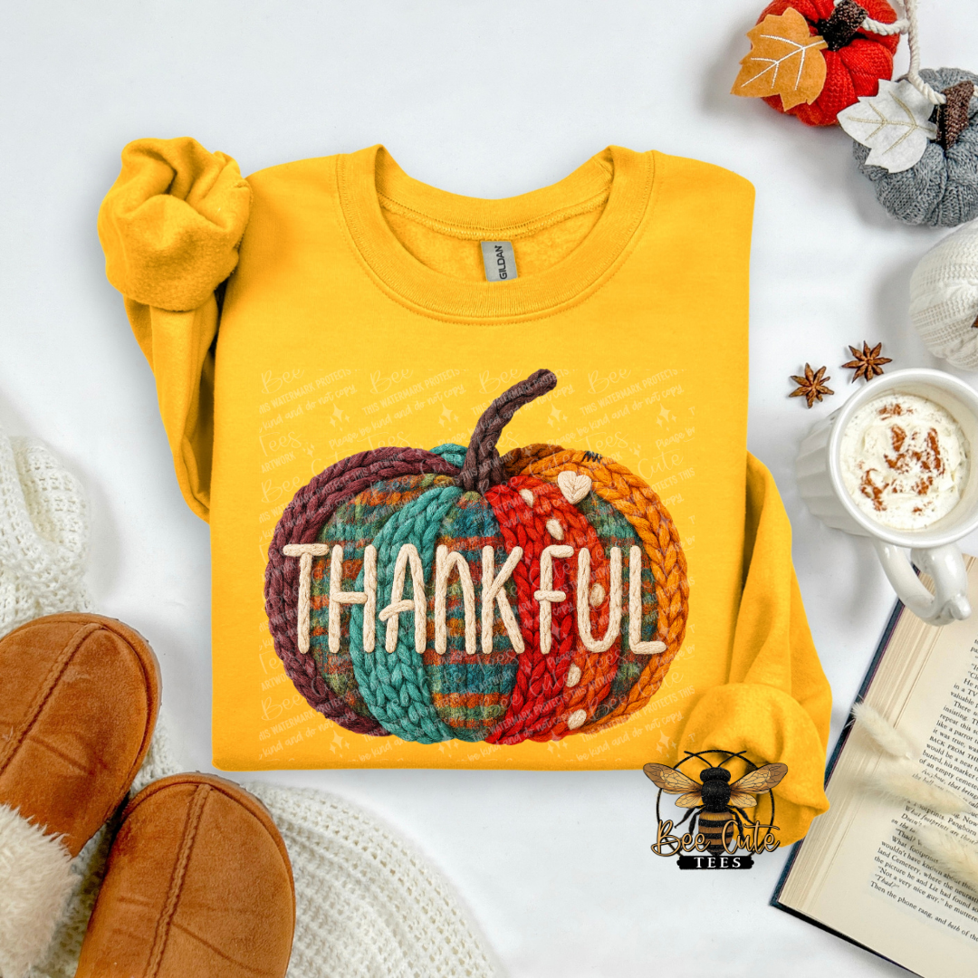 A folded yellow sweatshirt with a colorful autumn-themed 'THANKFUL' pumpkin graphic, styled in a cozy flat-lay arrangement with a mug of coffee, an open book, and brown slippers on a white background.