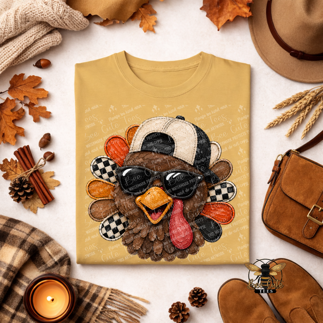 A folded tan T-shirt with a large embroidered turkey graphic wearing a black-and-white cap and dark sunglasses, surrounded by autumn props including leaves, a hat, shoes, a candle, and a small bag on a light surface.