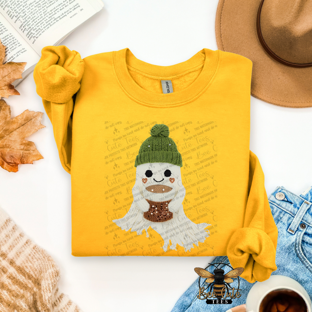 A folded yellow long-sleeve shirt with a cartoon ghost graphic wearing a green beanie and brown scarf, styled in a flat lay with a hat, jeans, leaves, a book, and a cup on a white background.