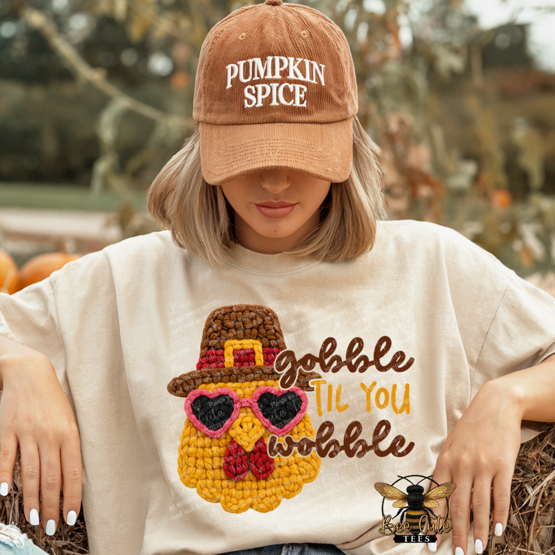 A woman wearing a tan baseball cap that says 'PUMPKIN SPICE' and a cream-colored t-shirt featuring a crocheted turkey graphic with the text 'Gobble Til You Wobble'. She is posed outdoors on a bed of straw with pumpkins and a blurred garden background.