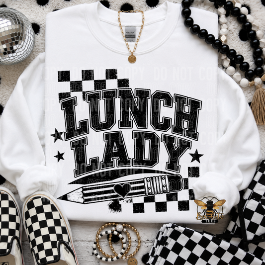 A white sweatshirt laid flat with a black-and-white checkerboard graphic reading “LUNCH LADY” and a pencil illustration. The sweatshirt is styled with black-and-white checkerboard shoes, beaded necklaces, a gold necklace, bracelets, and a matching checkered fabric piece on a textured white surface.