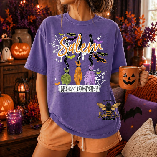 A woman wearing a purple graphic t-shirt with a Halloween-themed 'Salem Broom Company' design, holding an orange mug in a cozy, decorated autumn living room.