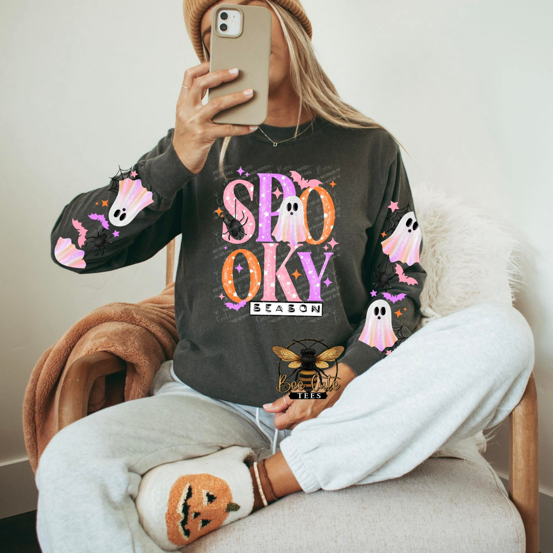 A woman takes a mirror selfie wearing a dark charcoal gray long-sleeve sweatshirt with a 'SPOOKY SEASON' graphic on the front, decorated with pink and purple ghosts and white pumpkins on the sleeves. She is sitting in a cozy armchair wearing light gray sweatpants and a pumpkin-themed sock.
