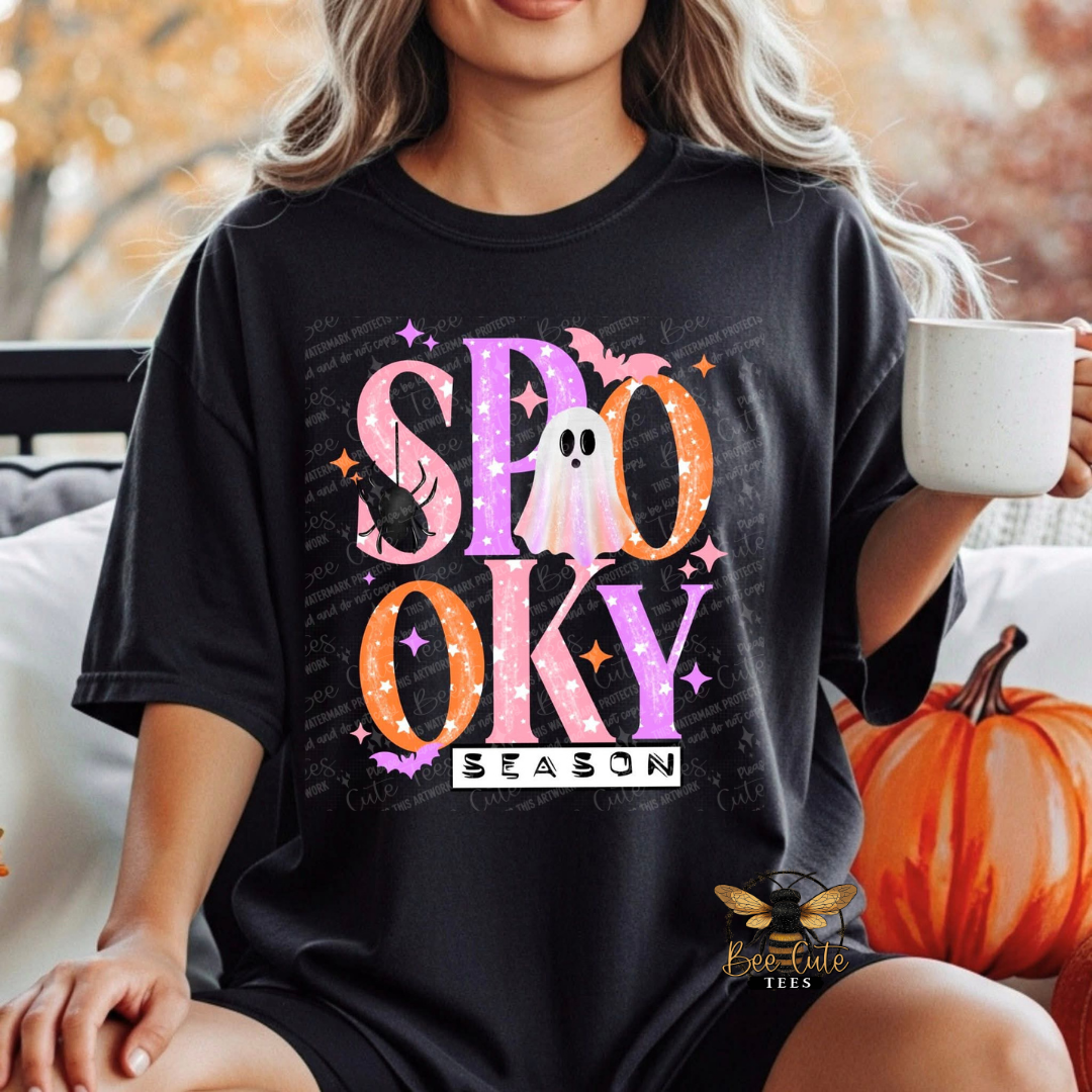 A woman wearing a black graphic t-shirt with a colorful 'SPOOKY SEASON' Halloween design, holding a white mug next to a pumpkin.