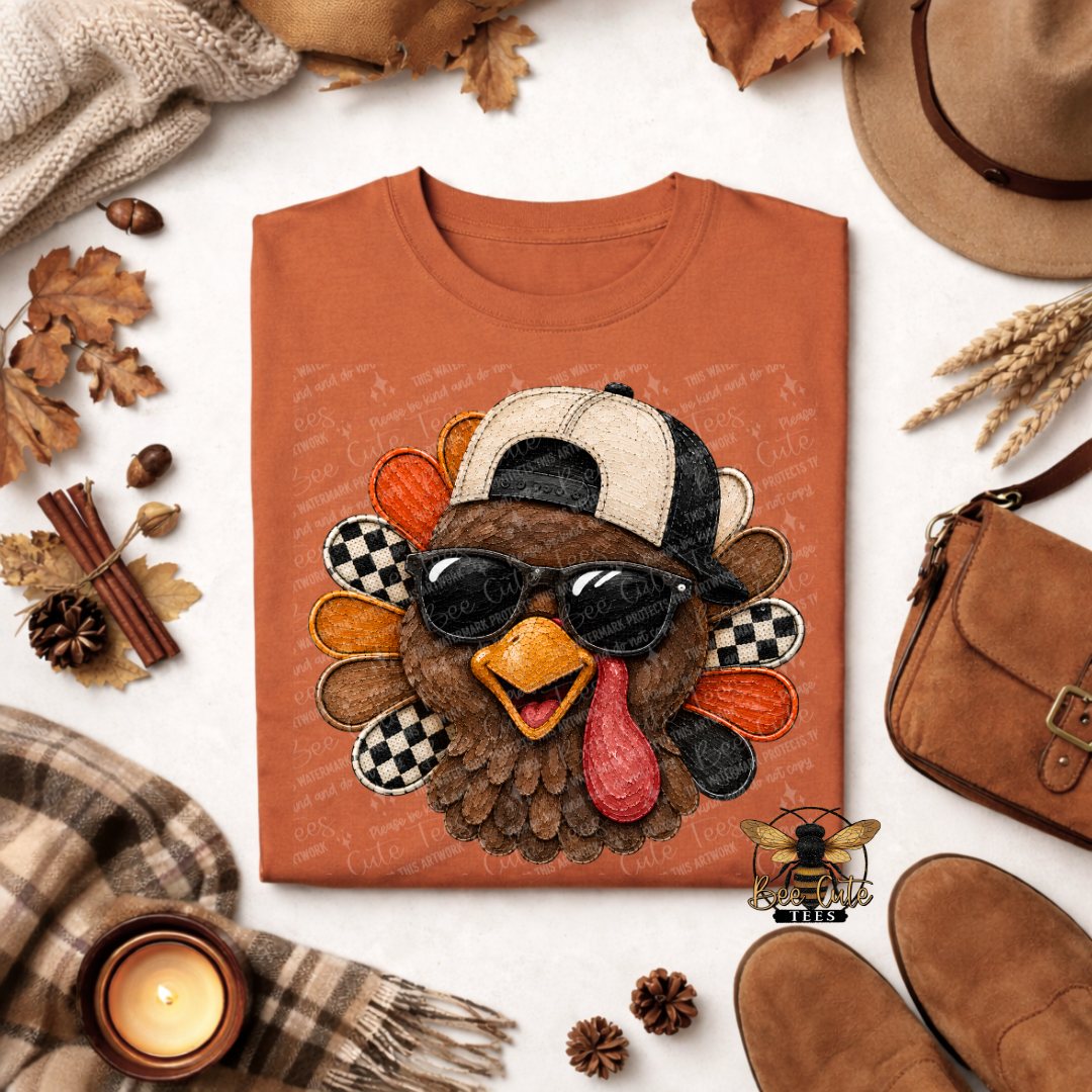 An orange crewneck sweatshirt laid flat on a light surface, surrounded by autumn props including leaves, a hat, a bag, a candle, and shoes. The sweatshirt features a large applique turkey design wearing a black-and-white cap and dark sunglasses.