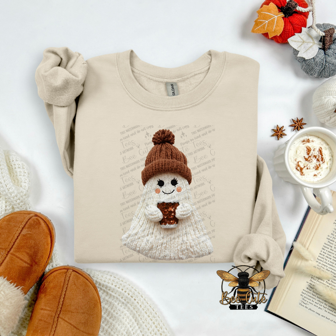 A beige long-sleeve sweatshirt laid flat on a white surface, featuring a cute knitted ghost graphic wearing a brown pom-pom hat and scarf. The sweatshirt is styled with cozy autumn props including a mug of hot cocoa, an open book, knit socks, and slippers.