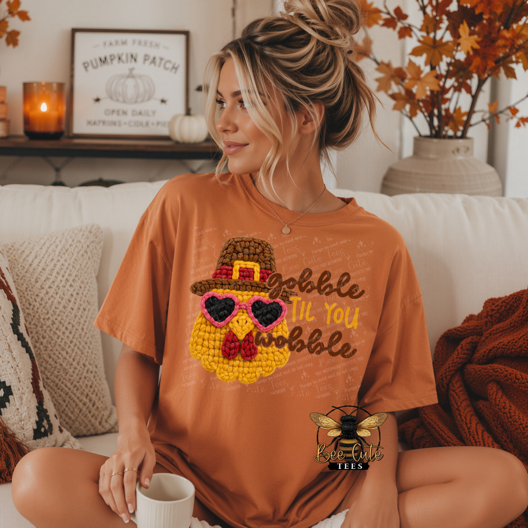A woman sits cross-legged on a white couch wearing an orange graphic t-shirt with a crocheted turkey design and the text 'Gobble Till You Wobble'. The background shows a cozy living room setting with autumn decorations.