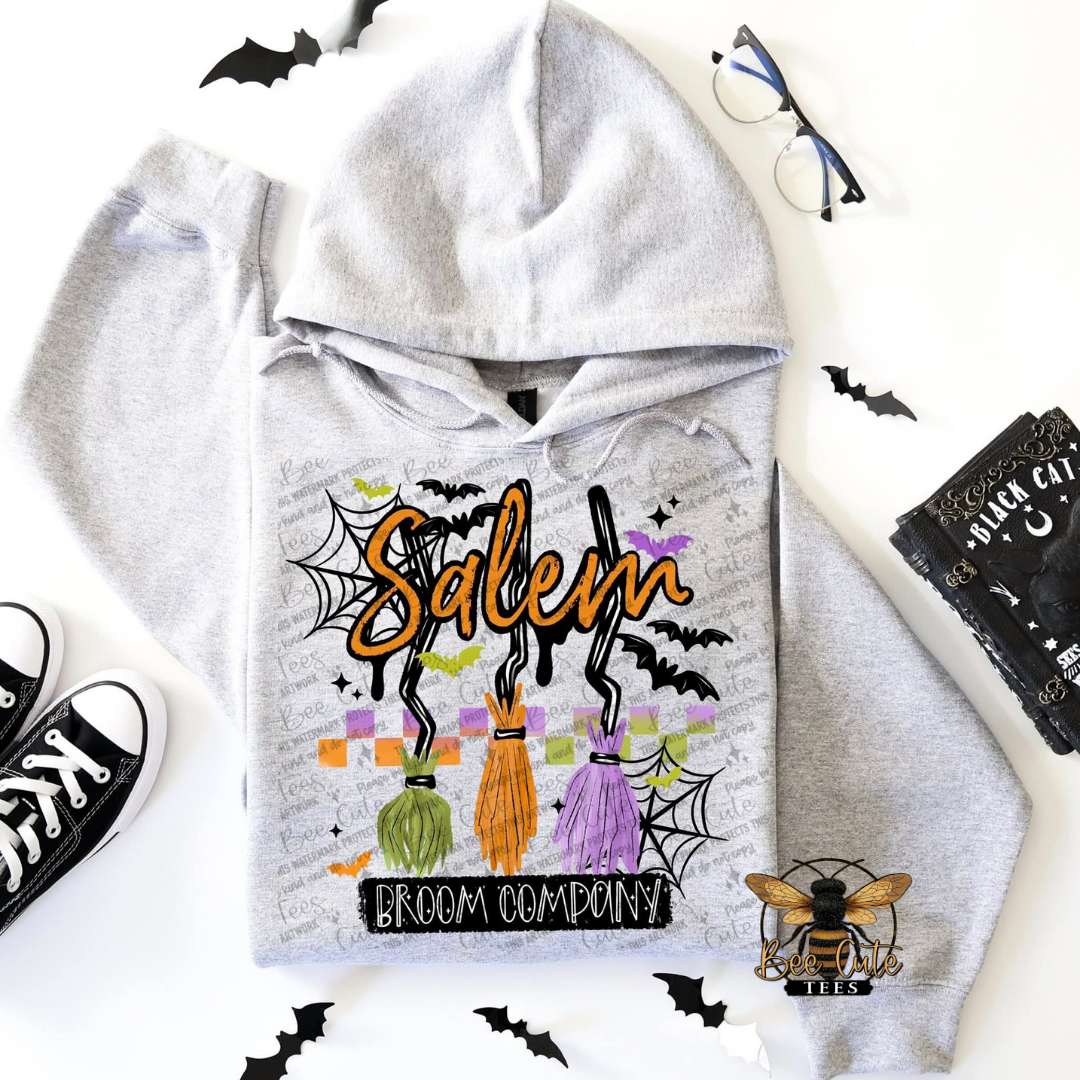 A light gray hoodie laid flat on a white background with Halloween-themed props around it, including black bat cutouts, glasses, black sneakers, a black card, and a bee logo. The hoodie has a large front print reading 'Salem' in orange with 'Broom Company' below, featuring three colorful brooms and spiderweb motifs.