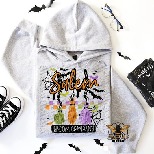 A light gray hoodie laid flat on a white background with Halloween-themed props around it, including black bat cutouts, glasses, black sneakers, a black card, and a bee logo. The hoodie has a large front print reading 'Salem' in orange with 'Broom Company' below, featuring three colorful brooms and spiderweb motifs.