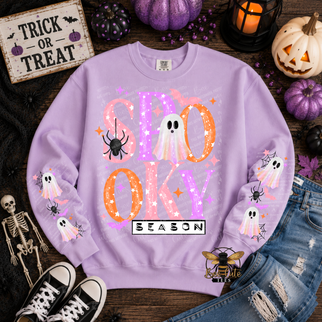 A lavender crewneck sweatshirt with a 'SPOOKY SEASON' Halloween graphic, styled in a flat lay with autumn props including pumpkins, a skeleton, jeans, and sneakers.