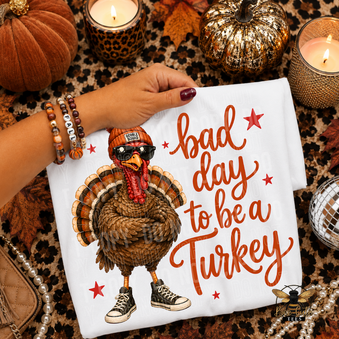 A folded white T-shirt with a cartoon turkey wearing sunglasses and sneakers and the phrase 'bad day to be a Turkey' in orange script, held by a person's hand in a fall-themed styled flat lay with pumpkins, candles, and autumn decor.