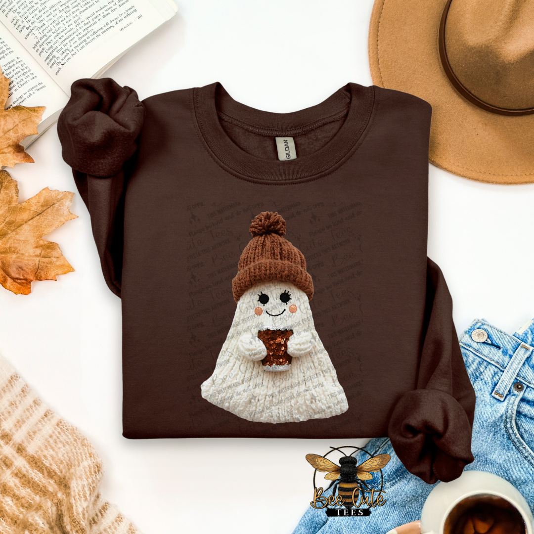 A dark brown long-sleeve t-shirt with a graphic of a cute white ghost wearing a brown knit hat and holding a mug, styled in an autumn flat-lay with a hat, jeans, leaves, and a cup of coffee.