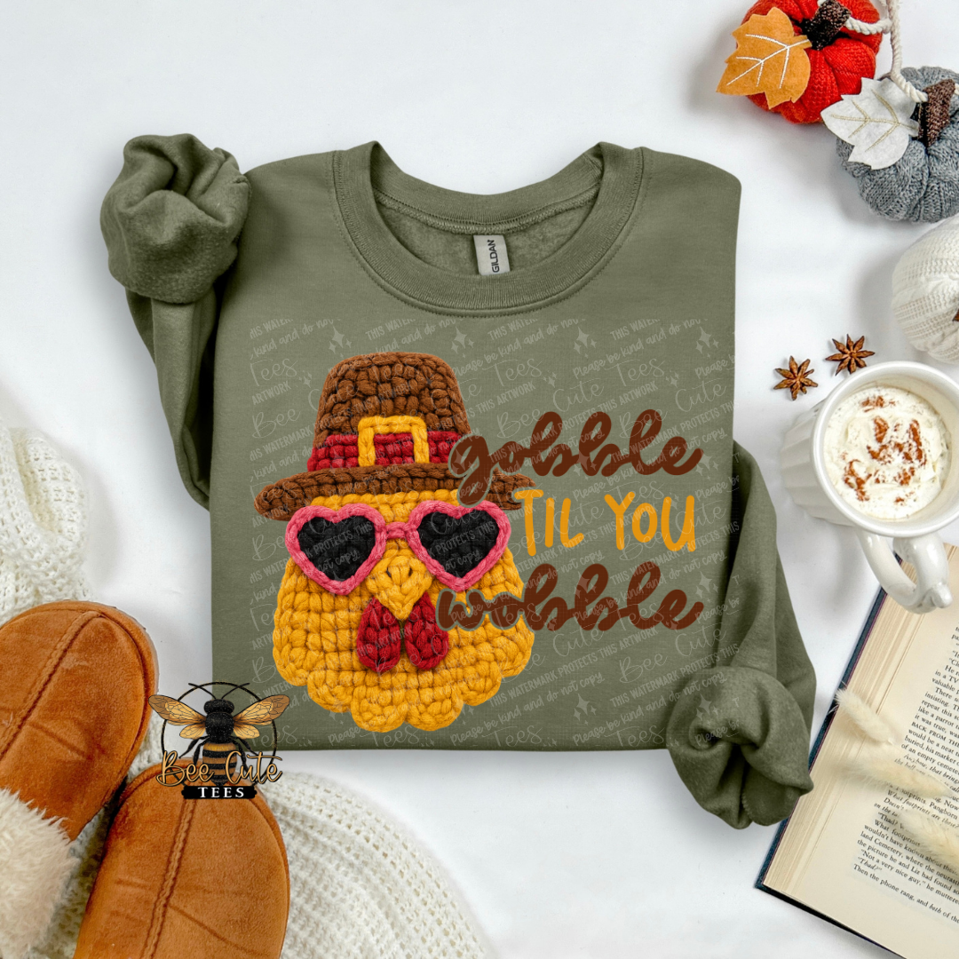 An olive green long-sleeve t-shirt with a crocheted turkey graphic wearing a brown hat and pink heart sunglasses, next to the text 'Gobble Til You Wobble'. The shirt is styled in a flat lay with autumn props including a mug of coffee, an open book, and a pair of brown slippers.