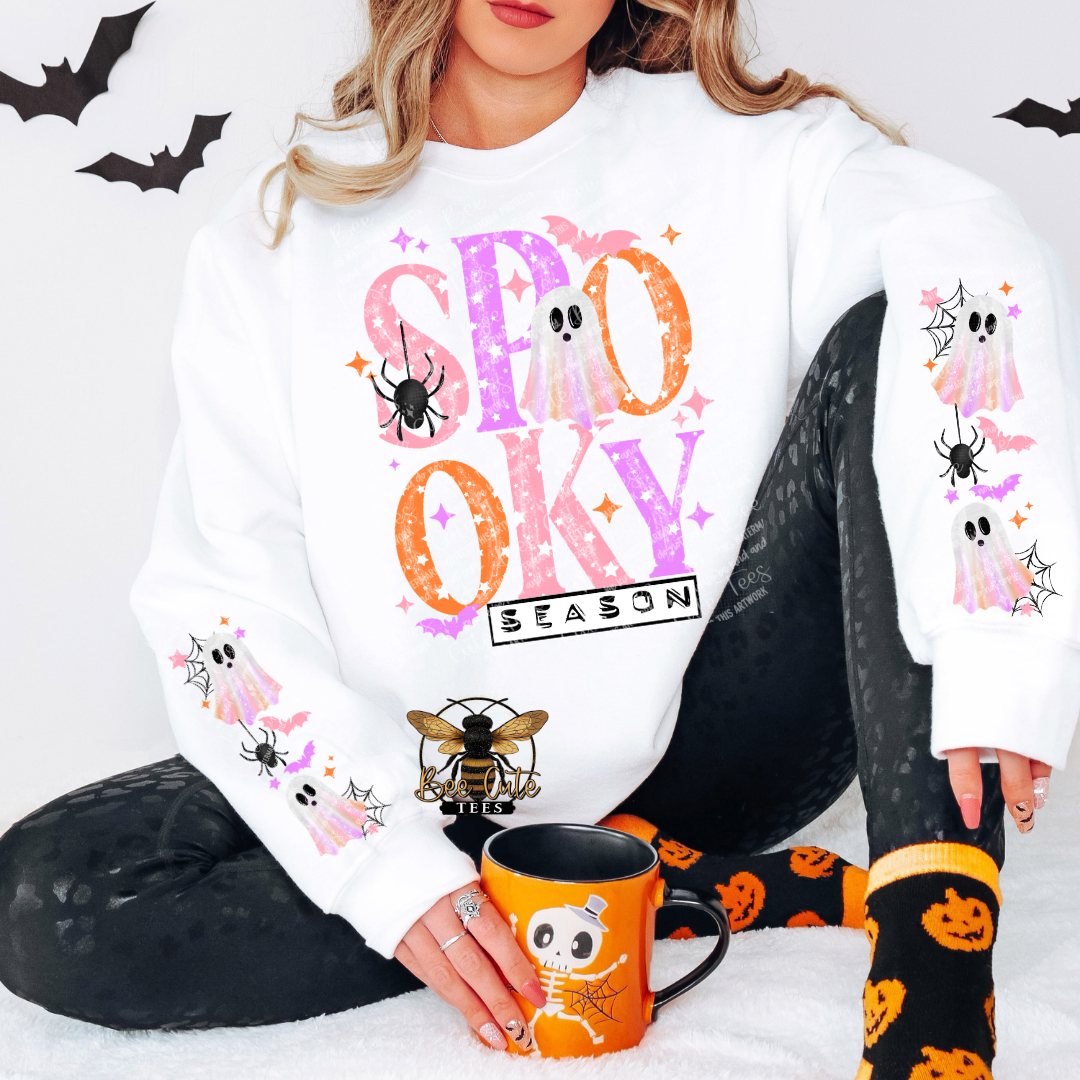 A woman wearing a white crewneck sweatshirt with a colorful 'SPOOKY SEASON' graphic on the front, styled with black leggings, orange and black patterned socks, and holding an orange mug with a skeleton design.