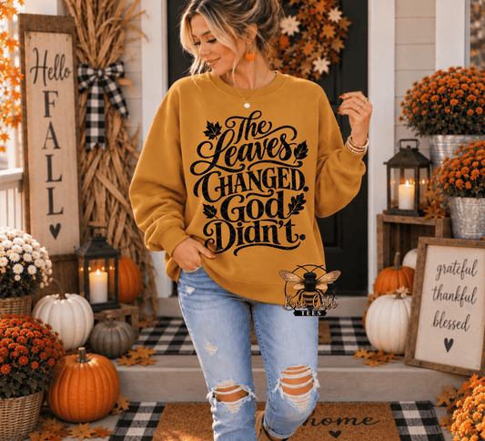 A woman wearing a mustard yellow crewneck sweatshirt with a black graphic text that reads 'The Leaves Changed, God Didn't.' stands on a decorated front porch with pumpkins, fall flowers, and autumn decorations.