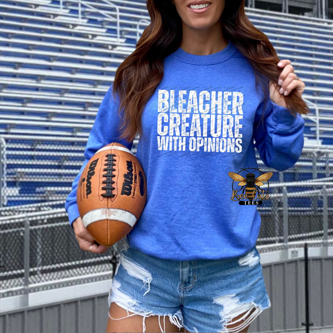 A woman wearing a royal blue sweatshirt with the text 'BLEACHER CREATURE WITH OPINIONS' in white, holding a football in front of stadium seating. A bee logo is visible on the lower right of the sweatshirt.