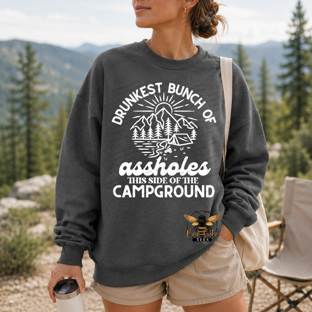 A woman wearing a dark gray crewneck sweatshirt with a white graphic that reads 'DRUNKEST BUNCH OF assholes THIS SIDE OF THE CAMPGROUND' and a small bee logo near the lower right front. She is outdoors in a mountain setting with trees and a camp chair visible.