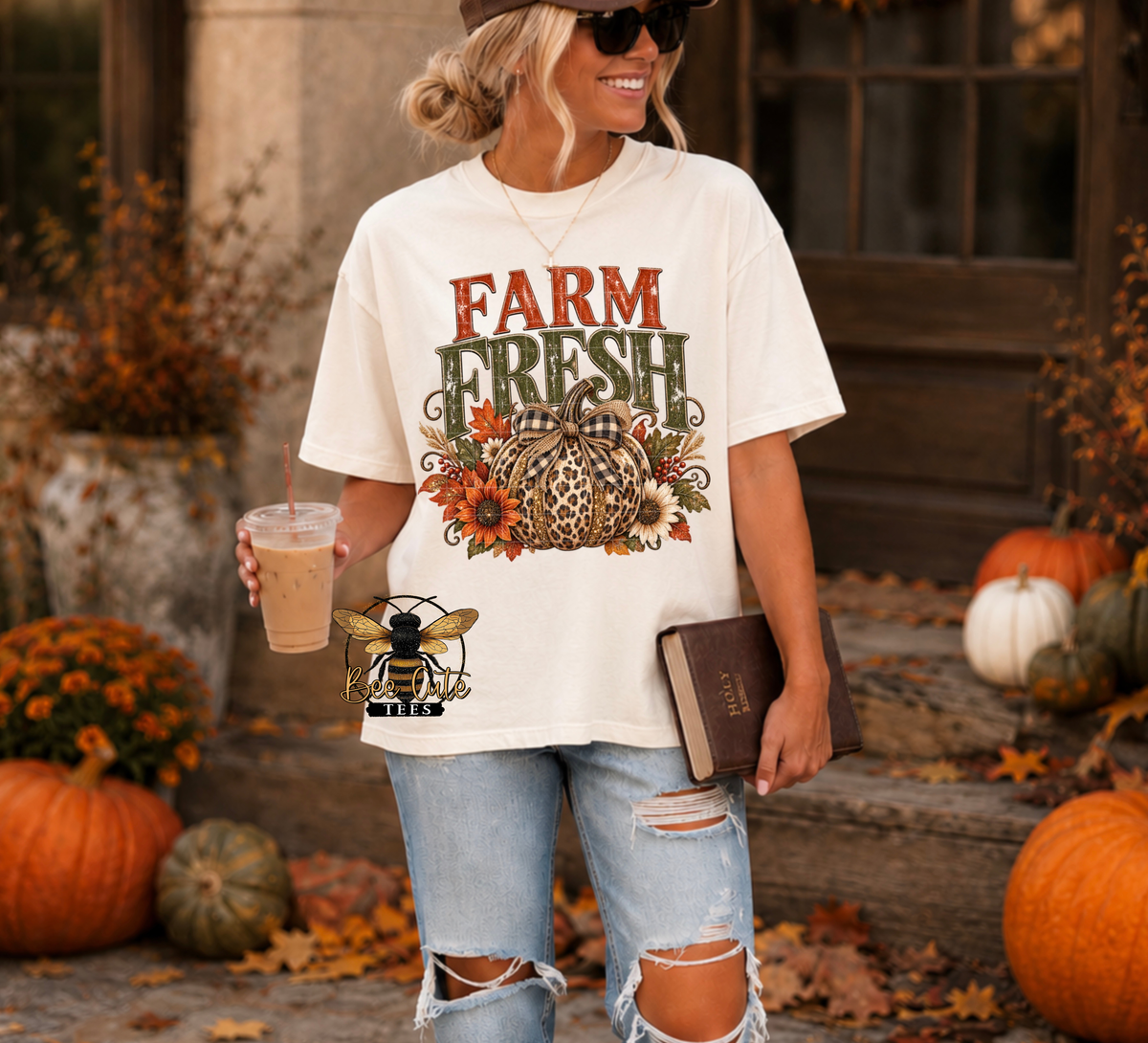 A woman wearing a graphic t-shirt with a 'FARM FRESH' autumn design, paired with ripped light-wash jeans, holding an iced coffee and a book in an outdoor fall-themed setting with pumpkins.
