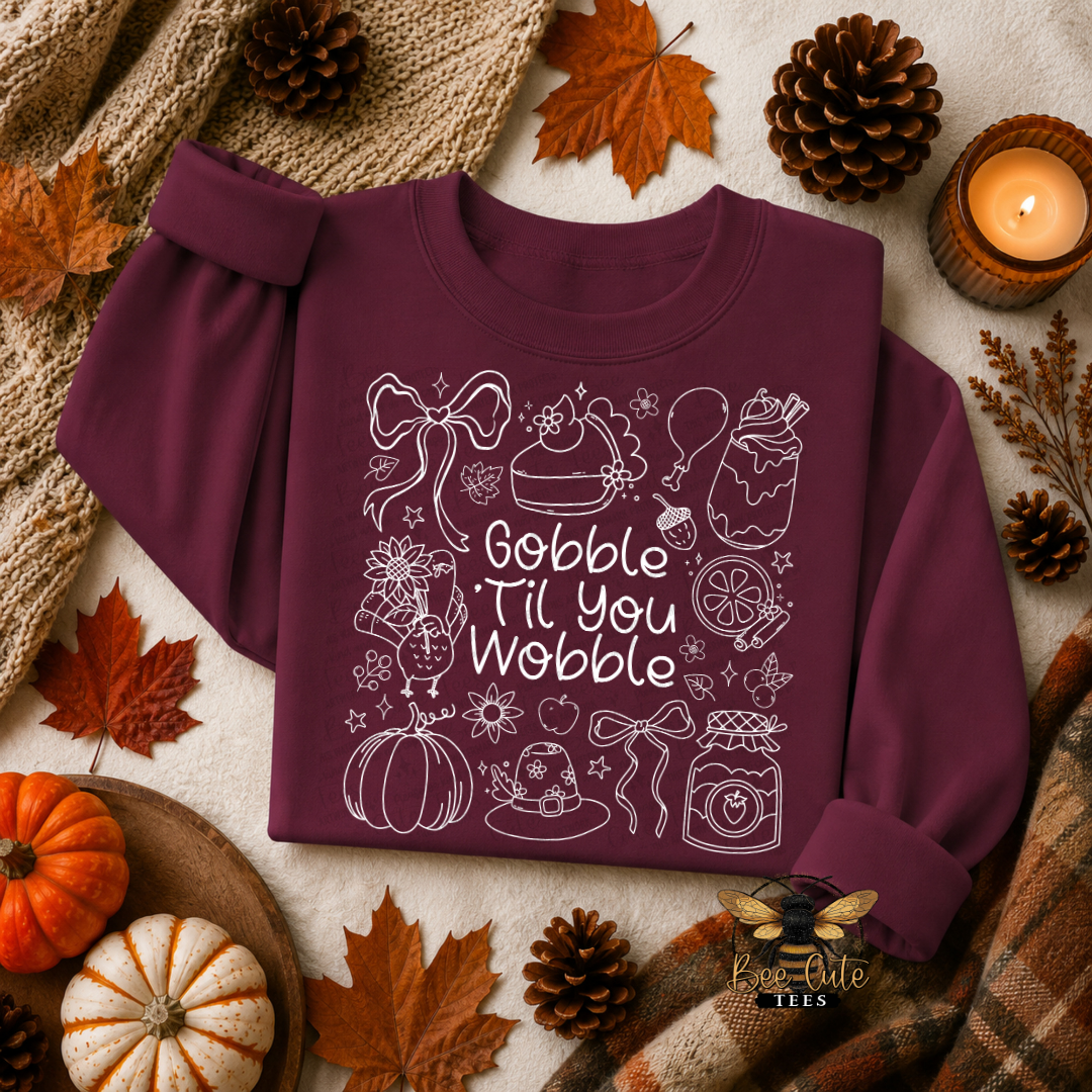 A folded long-sleeve crewneck sweatshirt in deep burgundy with white line-art illustrations and the phrase 'Gobble Til You Wobble' on the front, styled in a fall flat lay with pumpkins, pinecones, leaves, a candle, and plaid fabric.