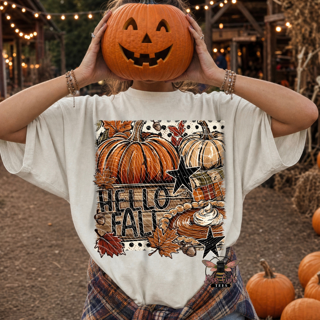 A person outdoors wearing a white graphic T-shirt with a large autumn-themed print reading 'HELLO FALL' and showing a carved orange pumpkin on the head, with pumpkins and a rustic fall setting in the background.