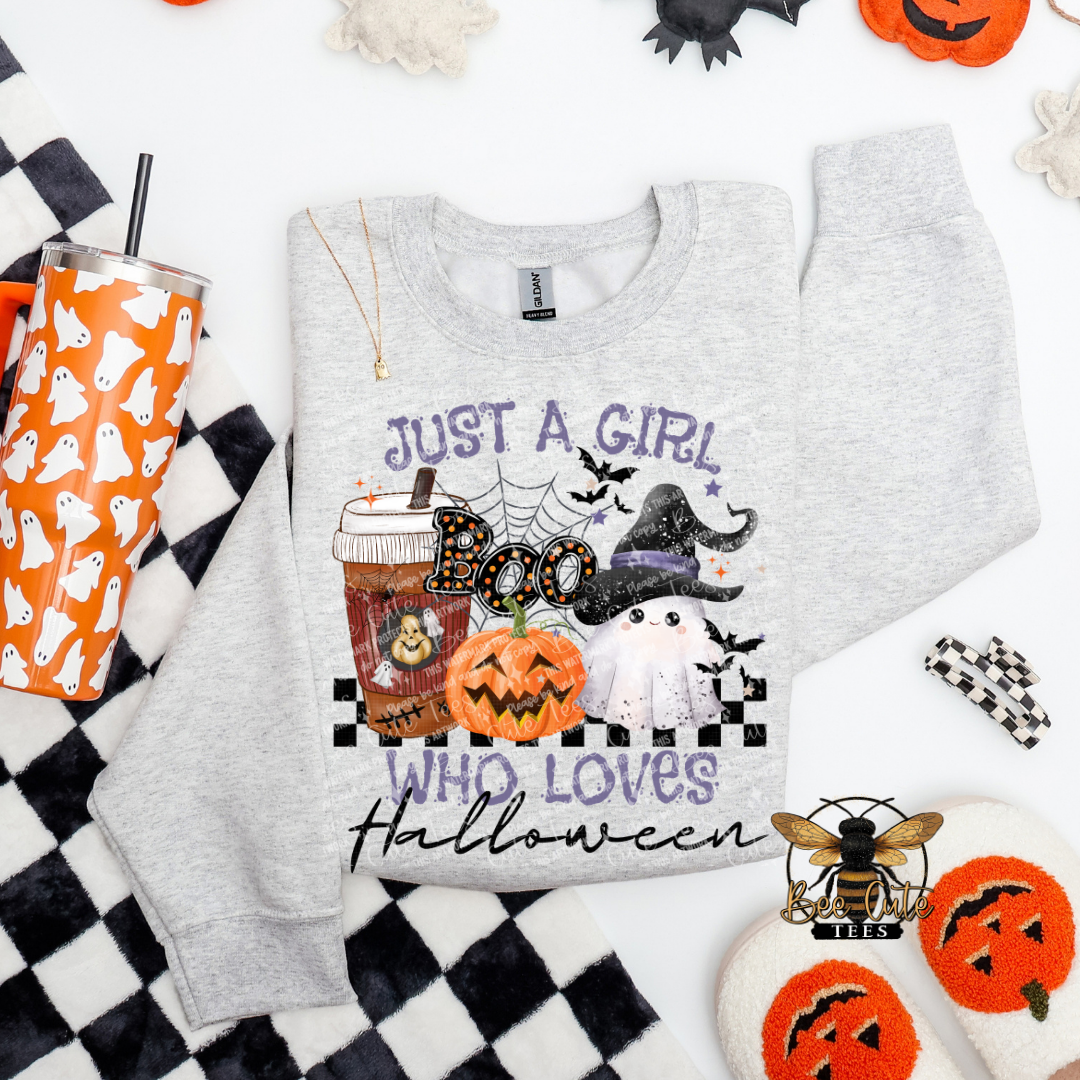 A light heather gray long-sleeve sweatshirt with a Halloween graphic print reading 'JUST A GIRL WHO LOVES Halloween' and featuring pumpkins, ghosts, bats, and a witch hat, styled in a flat lay with an orange ghost-patterned tumbler, black-and-white check fabric, and small decorative props.