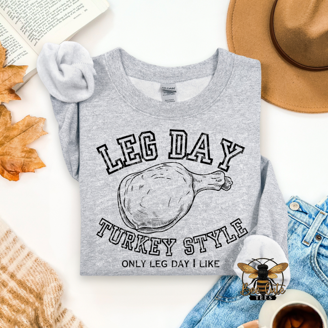 A folded heather gray graphic T-shirt is styled in a flat lay with autumn leaves, an open book, a tan hat, blue jeans, a knit sweater, and a cup of coffee. The shirt features a turkey leg illustration and the text 'LEG DAY TURKEY STYLE' and 'ONLY LEG DAY I LIKE'.