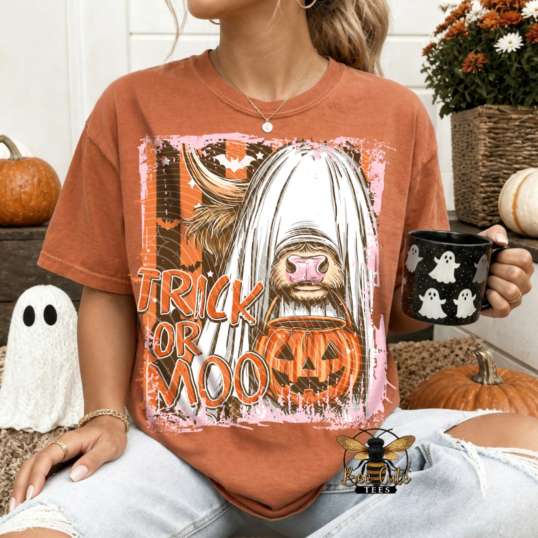 A woman wearing a rust-orange graphic t-shirt with a Halloween design featuring a ghosted cat and the text 'TRICK OR MOO'. She is holding a black mug with white ghosts, sitting in a cozy, autumn-themed setting with pumpkins and a ghost decoration.