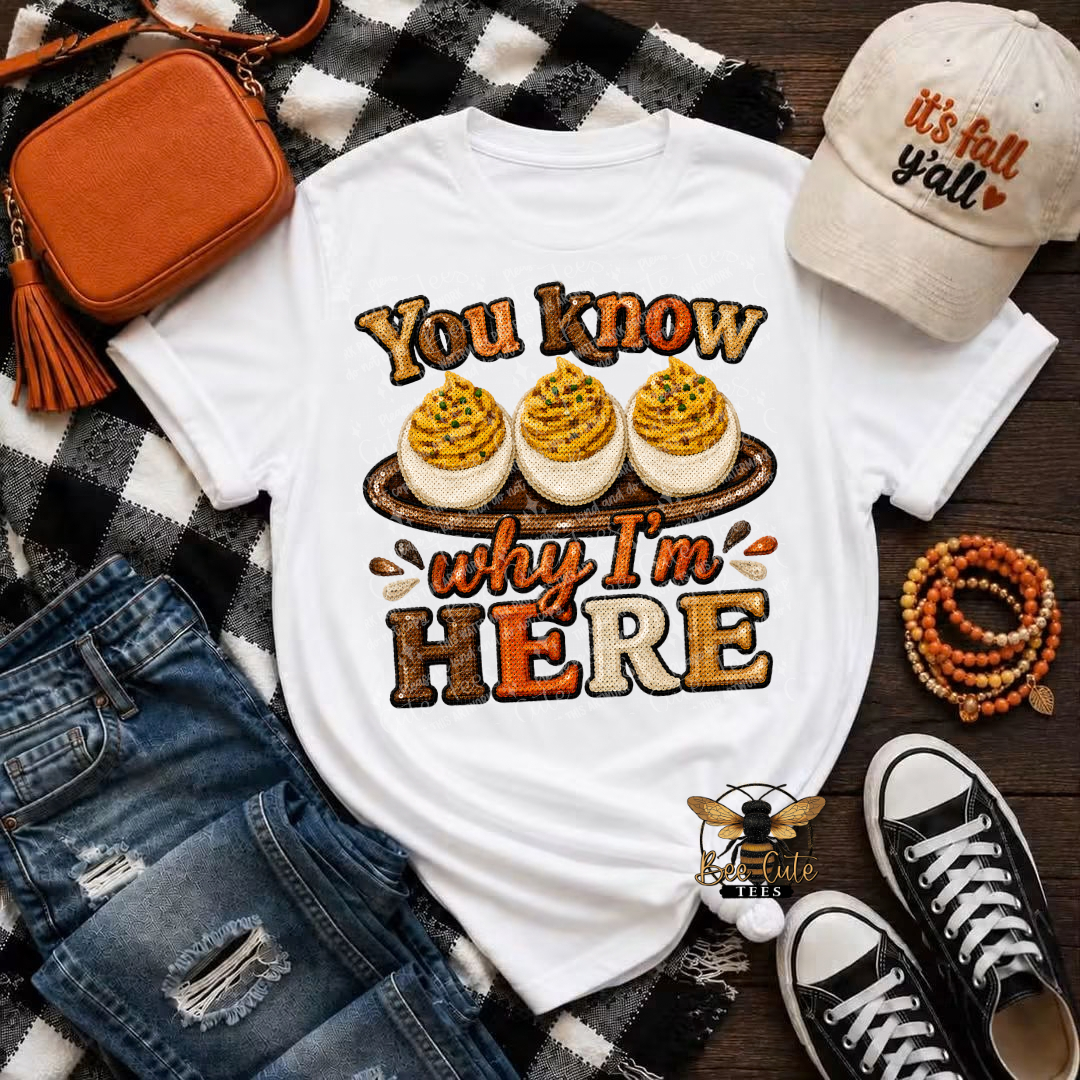 A white graphic T-shirt laid flat on a black-and-white plaid blanket, surrounded by styled fall accessories including an orange tassel crossbody bag, a beige baseball cap reading 'it's fall y'all', distressed blue jeans, a beaded bracelet, and black low-top sneakers. The shirt graphic shows three deviled eggs with the text 'You know why I'm HERE'.