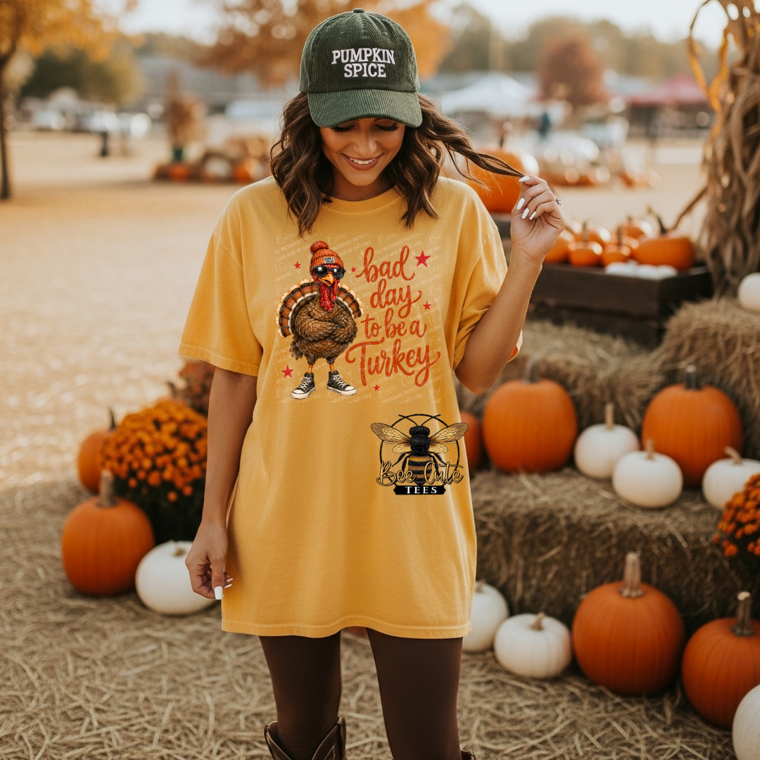 A woman stands outdoors in a fall-themed pumpkin patch wearing a mustard yellow graphic t-shirt with a turkey and bee logo, a green 'Pumpkin Spice' cap, and brown leggings. The background features stacked hay bales, pumpkins, and autumn foliage.