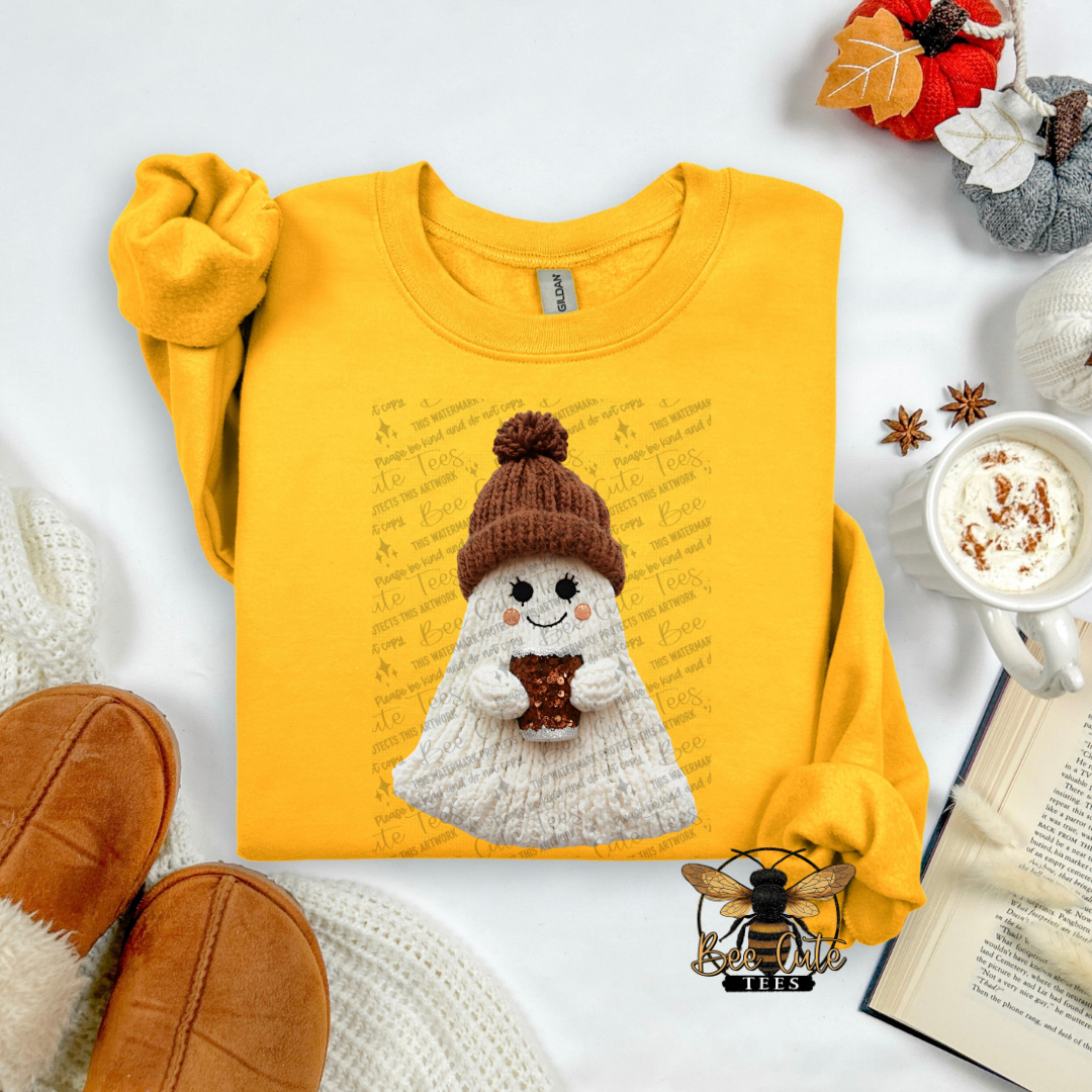 A folded yellow sweatshirt with a knitted ghost graphic holding a hot cocoa, styled in a cozy flat-lay with brown slippers, a mug of cocoa, an open book, and autumn leaves on a white background.