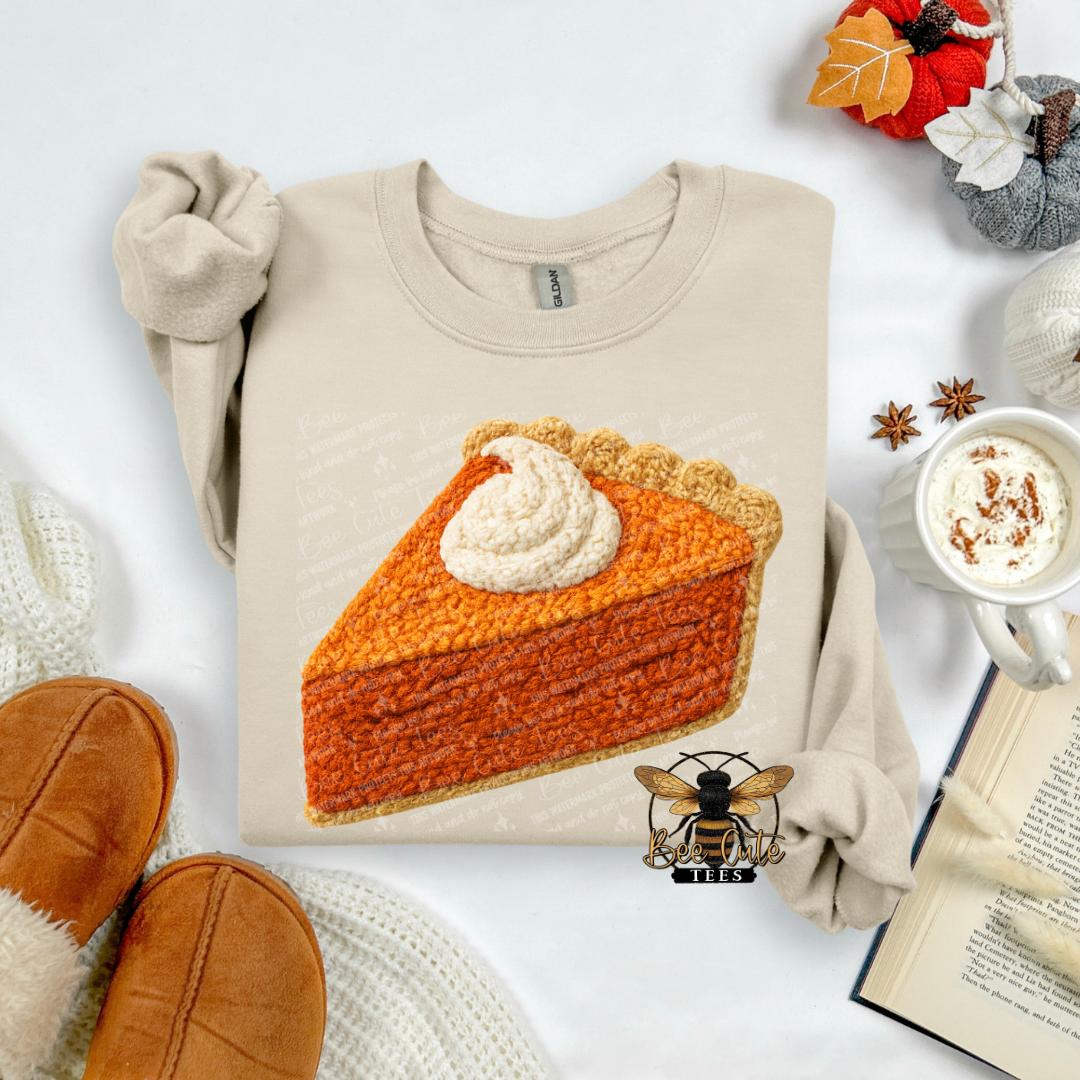 A beige long-sleeve sweatshirt laid flat on a white surface, featuring a large printed illustration of a pumpkin pie slice with whipped cream. The sweatshirt is styled with autumn props including tan slippers, a mug of hot cocoa, an open book, and fall leaves.
