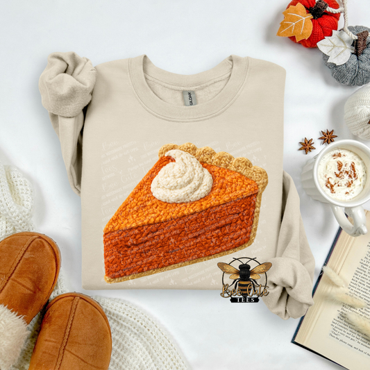 A beige long-sleeve sweatshirt laid flat on a white surface, featuring a large printed illustration of a pumpkin pie slice with whipped cream. The sweatshirt is styled with autumn props including tan slippers, a mug of hot cocoa, an open book, and fall leaves.