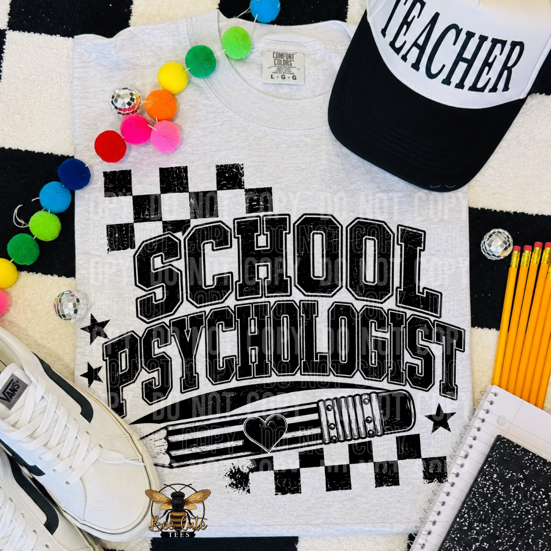 A white graphic T-shirt laid flat with bold black text reading 'SCHOOL PSYCHOLOGIST' and a clipboard/heart design. The shirt is styled with a black-and-white cap, colorful pom-poms, pencils, a sneaker, and a notebook on a black-and-white checkered surface.