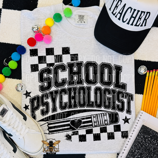 A white graphic T-shirt laid flat with bold black text reading 'SCHOOL PSYCHOLOGIST' and a clipboard/heart design. The shirt is styled with a black-and-white cap, colorful pom-poms, pencils, a sneaker, and a notebook on a black-and-white checkered surface.