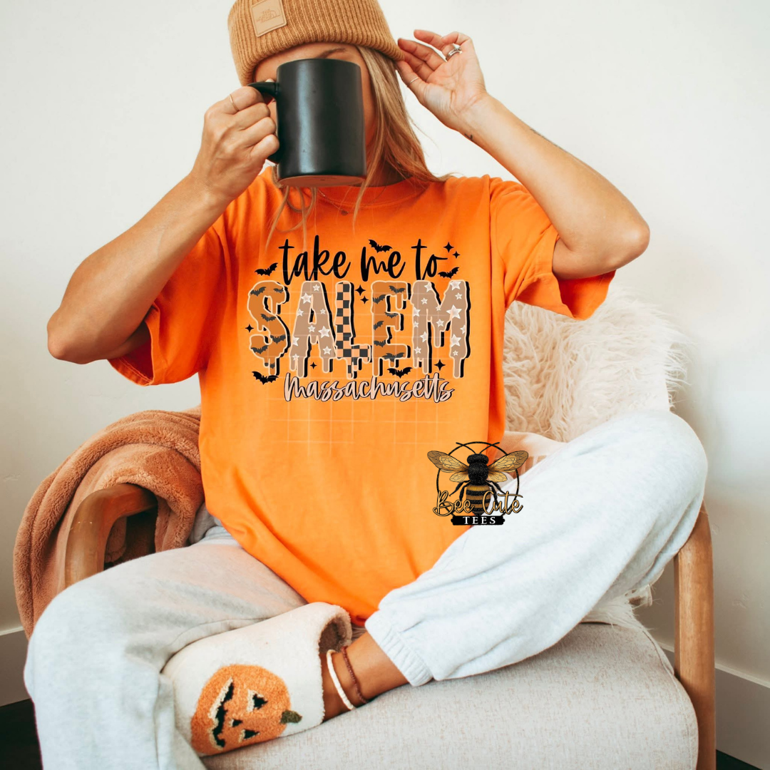 A woman sitting in a chair wearing an orange graphic T-shirt that reads 'take me to SALEM Massachusetts' with a bee logo, paired with white sweatpants and a tan beanie while holding a black mug.