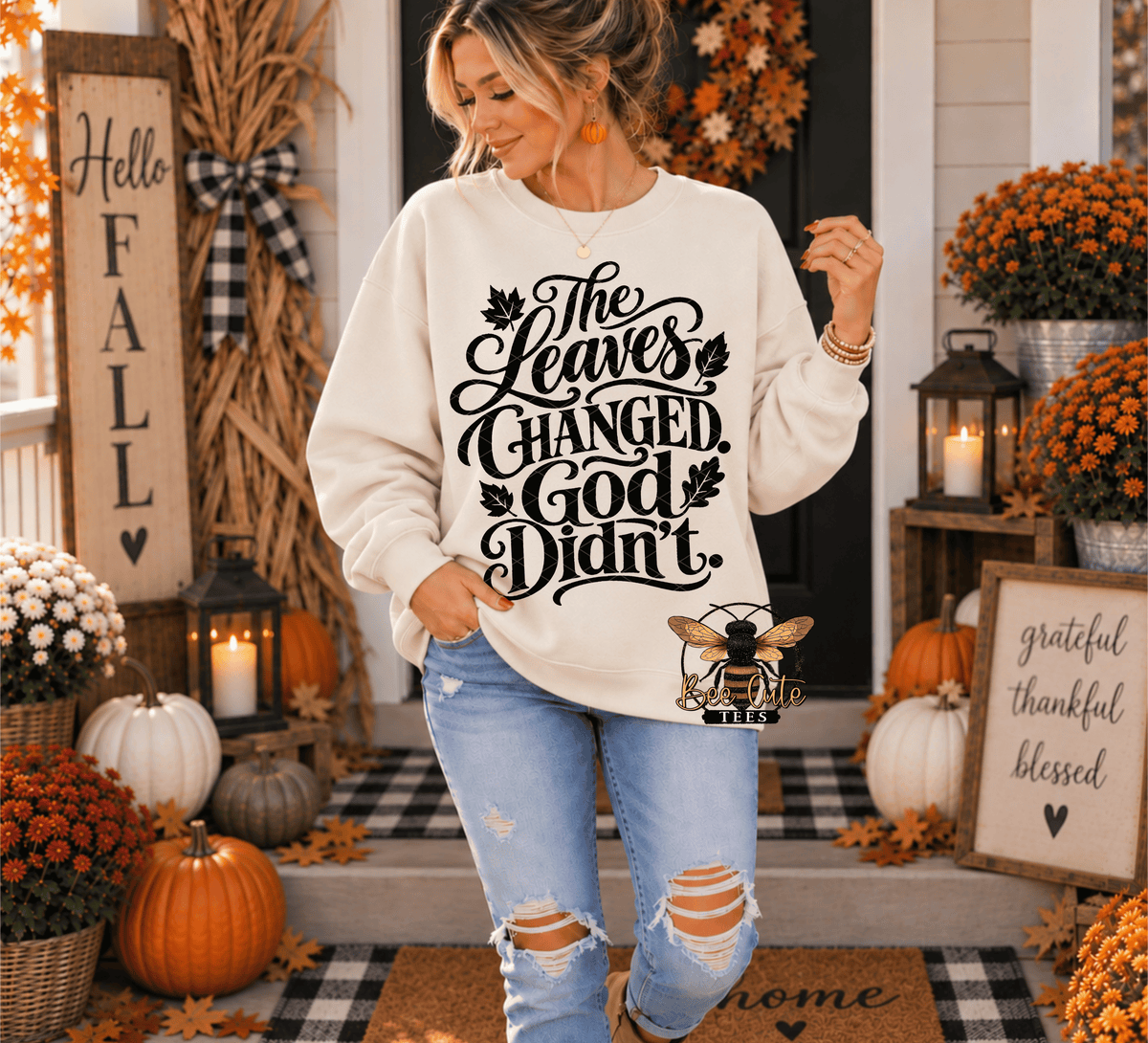 A woman wearing a cream-colored crewneck sweatshirt with a black graphic that reads 'The Leaves Changed, God Didn't.' stands on a decorated fall porch with pumpkins, mums, and autumn decorations.