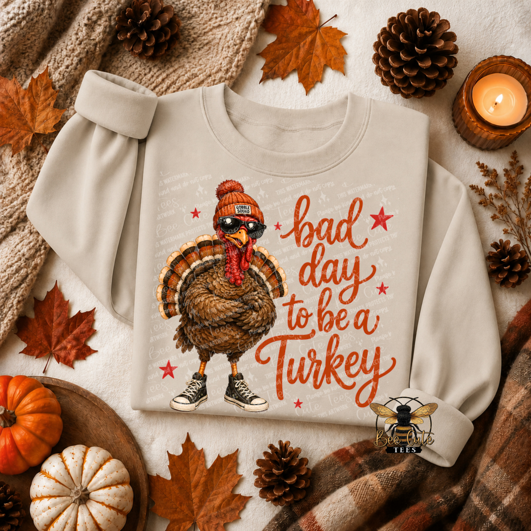A light beige crewneck sweatshirt laid flat on a cozy autumn-themed background. The sweatshirt features a cartoon turkey wearing a beanie and sunglasses next to the text 'bad day to be a Turkey' in orange.