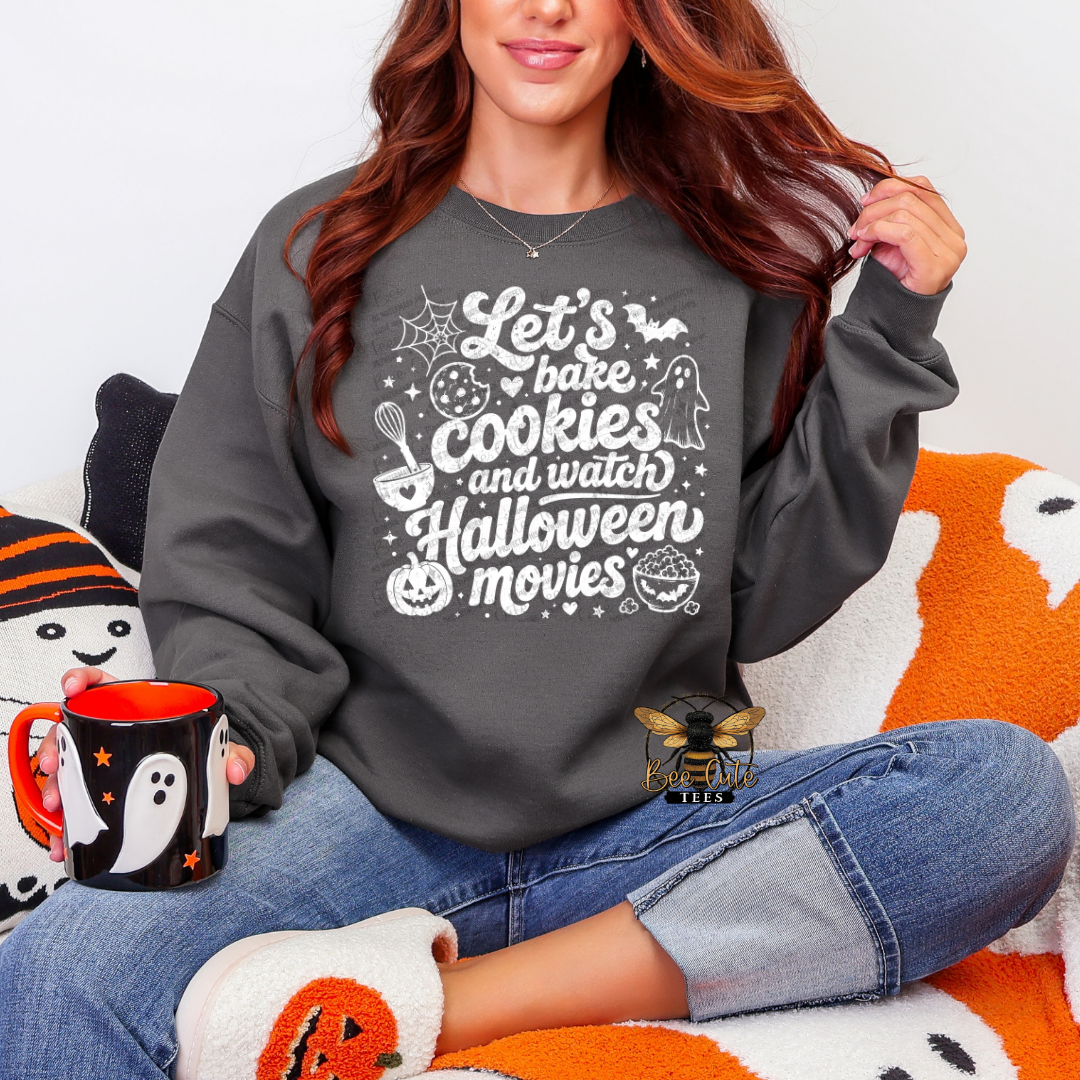 A woman wearing a dark gray crewneck sweatshirt with a white 'Let's bake cookies and watch Halloween movies' graphic, sitting on an orange and white blanket and holding a black mug with a ghost design.