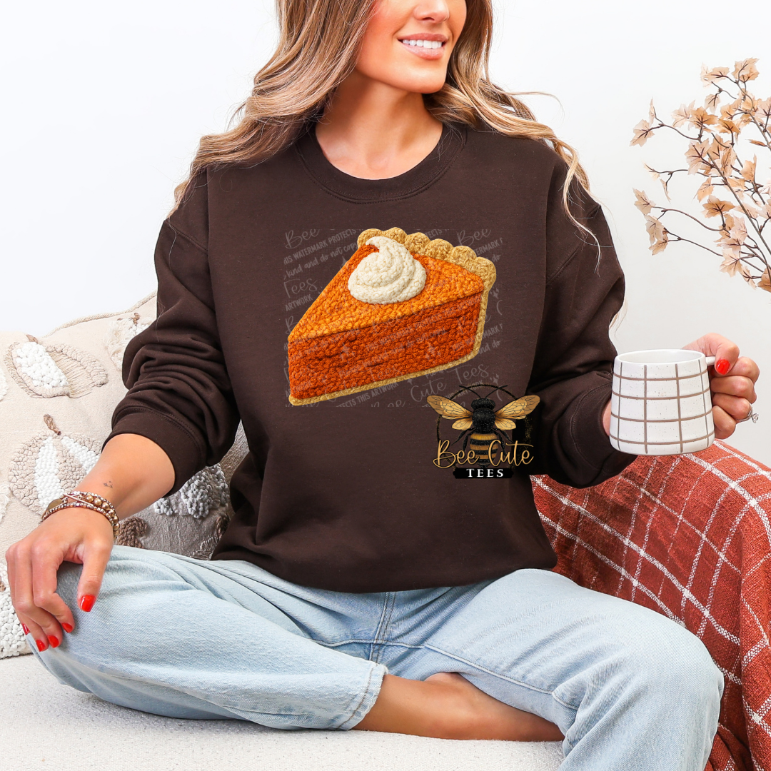A woman sitting indoors wearing a dark brown sweatshirt with a large pumpkin pie graphic and a bee logo that reads 'Bee Cute Tees'. She is holding a white mug with a brown grid pattern.